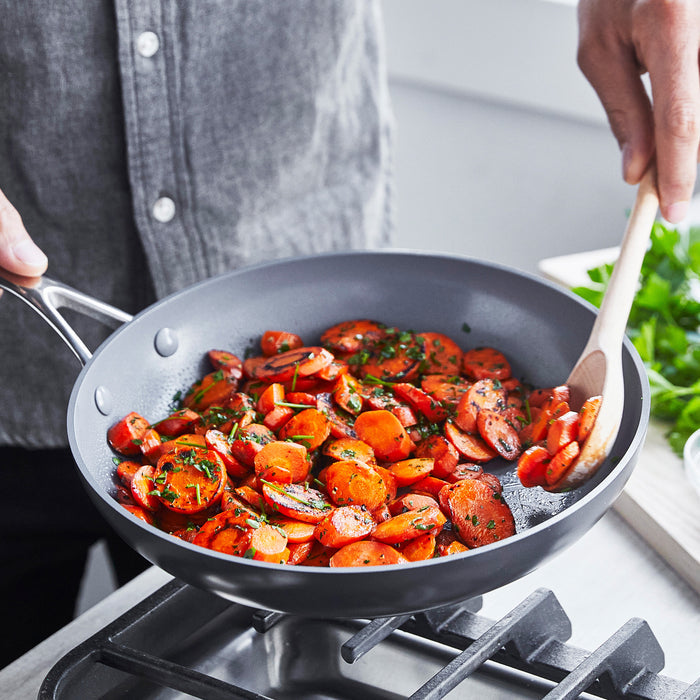 Carrots sautéing with herbs in a gray ceramic 12-inch frypan, stirred with a wooden spoon.