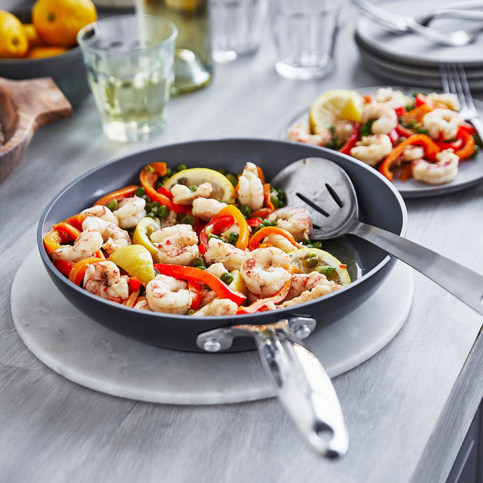 Ceramic nonstick skillet with shrimp, red peppers, and lemon slices on a table.
