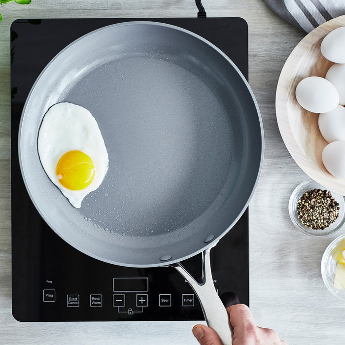 Hand holding a ceramic nonstick frypan with a sunny-side up egg on an induction cooktop, eggs next to the pan.