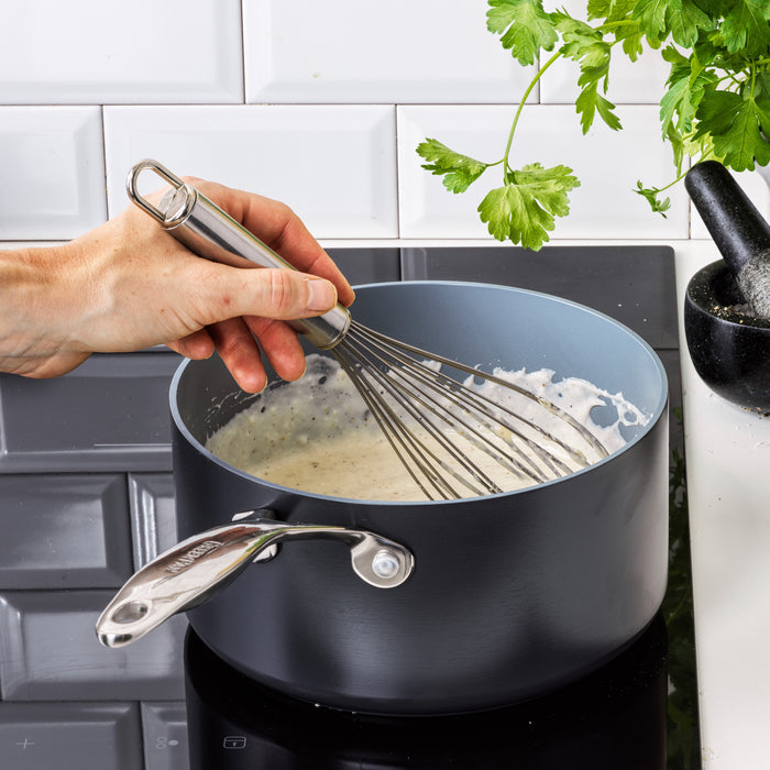 A hand stirs a creamy white sauce with a metal whisk in the Valencia Pro Ceramic Nonstick 1.5-Quart Saucepan on a stovetop, with fresh parsley and a mortar and pestle nearby.