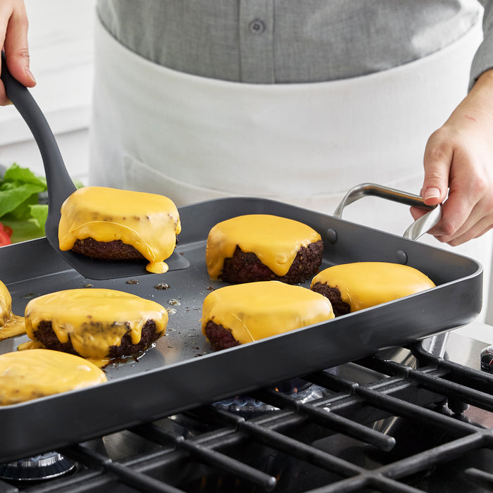 Cheeseburgers being cooked and flipped with a spatula on a large ceramic nonstick double burner griddle.