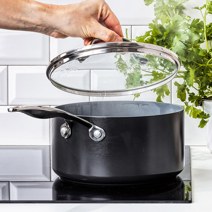 Hand lifting lid from steaming 2-quart ceramic nonstick saucepan on stove, fresh herbs visible in the background.