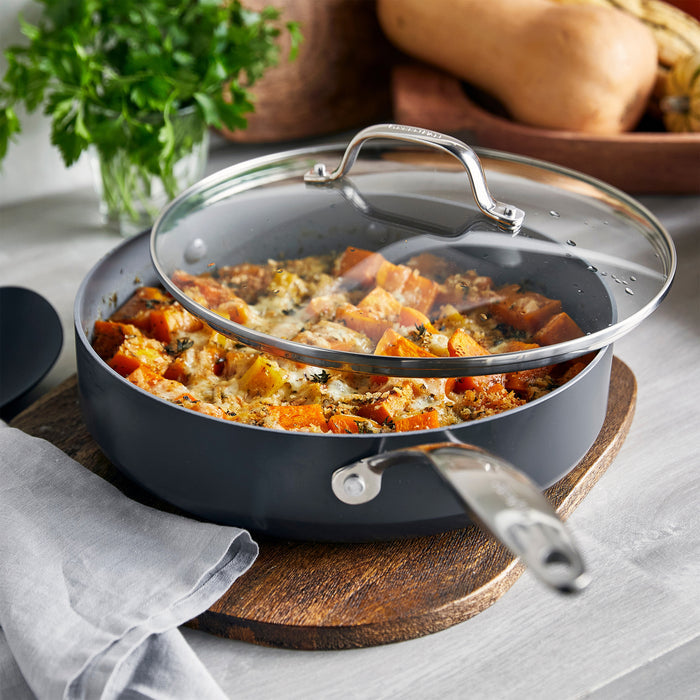 Ceramic sauté pan with lid filled with roasted vegetables, next to a board with fresh herbs.