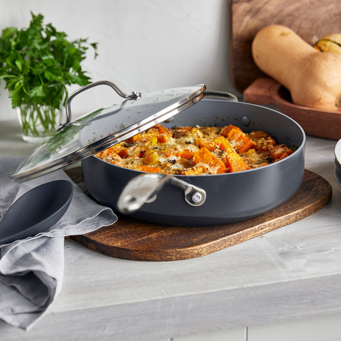 Gray ceramic sauté pan with lid on a wooden board, next to fresh herbs and yellow squash.