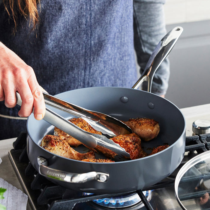 Chicken being cooked with tongs in a grey ceramic sauté pan on a gas stove.
