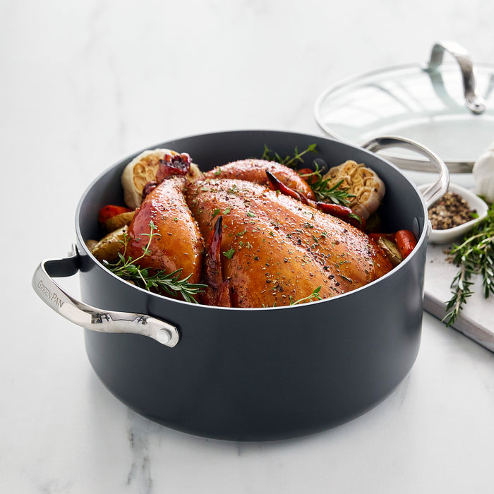 A whole roasted chicken with herbs and vegetables sits in the Valencia Pro Ceramic Nonstick 5.5-Quart Stockpot with Lid on a white countertop, with seasonings nearby.