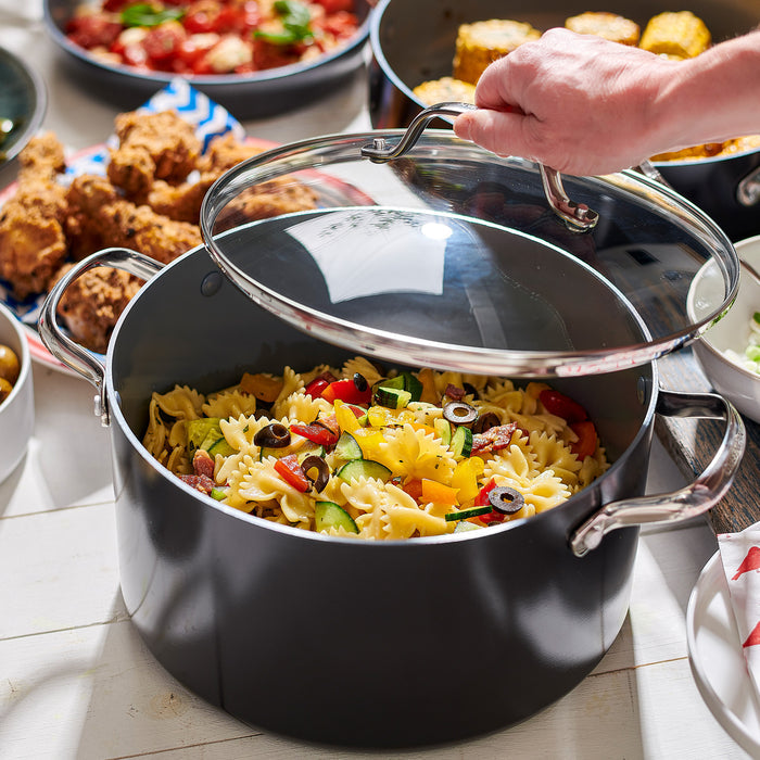 A hand lifts the lid of a Valencia Pro Ceramic Nonstick 5.5-Quart Stockpot filled with bowtie pasta, vegetables, and olives. In the background, other dishes like fried chicken and corn are visible on the table.