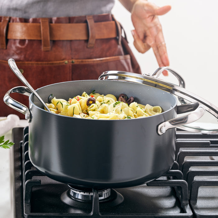 A pot of cooked pasta with olives and herbs sits on a gas stove, nestled in a Valencia Pro Ceramic Nonstick 5.5-Quart Stockpot with Lid. A person in a brown apron stands behind it, holding the lid and gesturing.