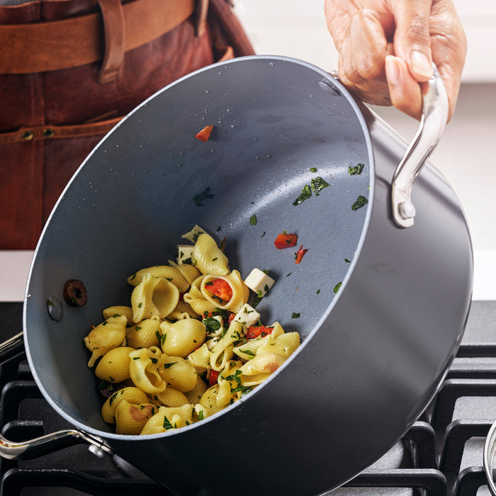 A person in a brown apron tilts a Valencia Pro Ceramic Nonstick 5.5-Quart Stockpot with Lid over a stove, revealing cooked shell pasta mixed with chopped vegetables and herbs inside.