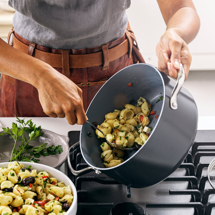 A person pours freshly cooked pasta with vegetables from a Valencia Pro Ceramic Nonstick 5.5-Quart Stockpot with Lid into a serving bowl on the stove, with parsley on the counter and a gray plate nearby.