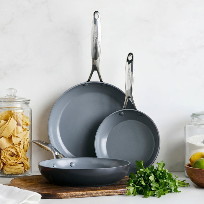 The Valencia Pro Ceramic Nonstick 8", 9.5", and 11" Frypan Set in gray, with stainless steel handles, sits on a wooden cutting board beside jars of pasta, lemons, and fresh parsley, highlighting their durable and scratch-resistant design.