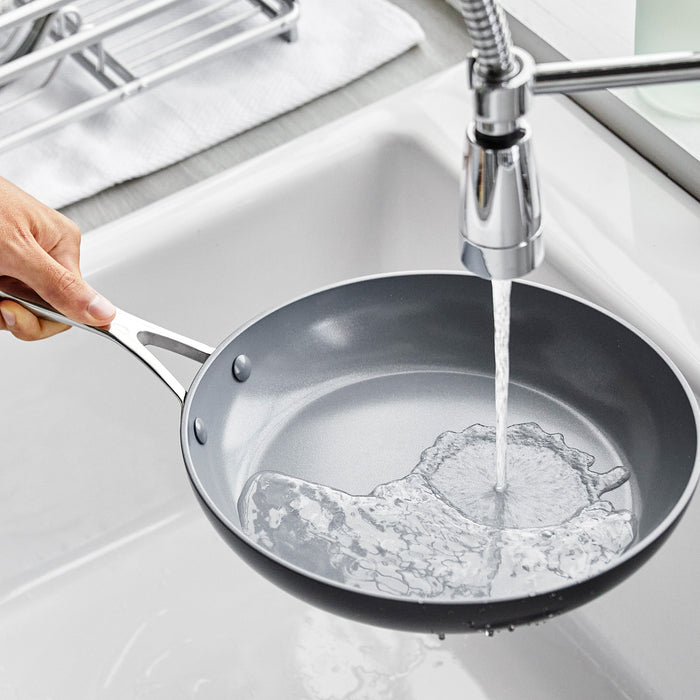 A hand rinsing an 8-inch ceramic nonstick frypan under running water in a kitchen sink.