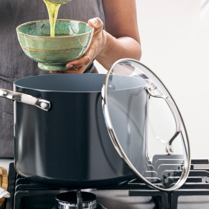 Green liquid being poured from a bowl into a gray ceramic stockpot on the stove, lid resting nearby.